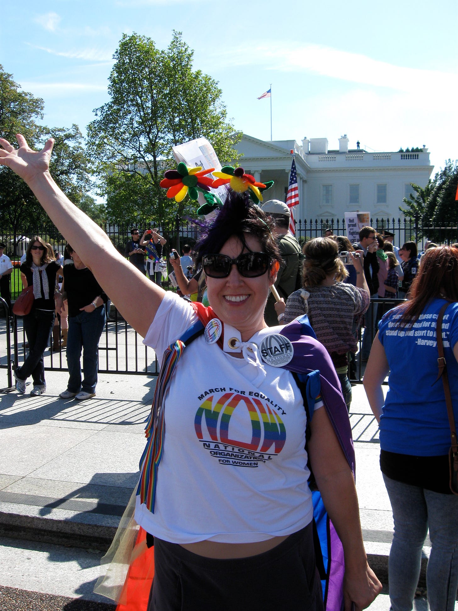 A photo of the author wearing pride clothing gesturing toward the sky with her hand and smiling broadly in front of the US Capital