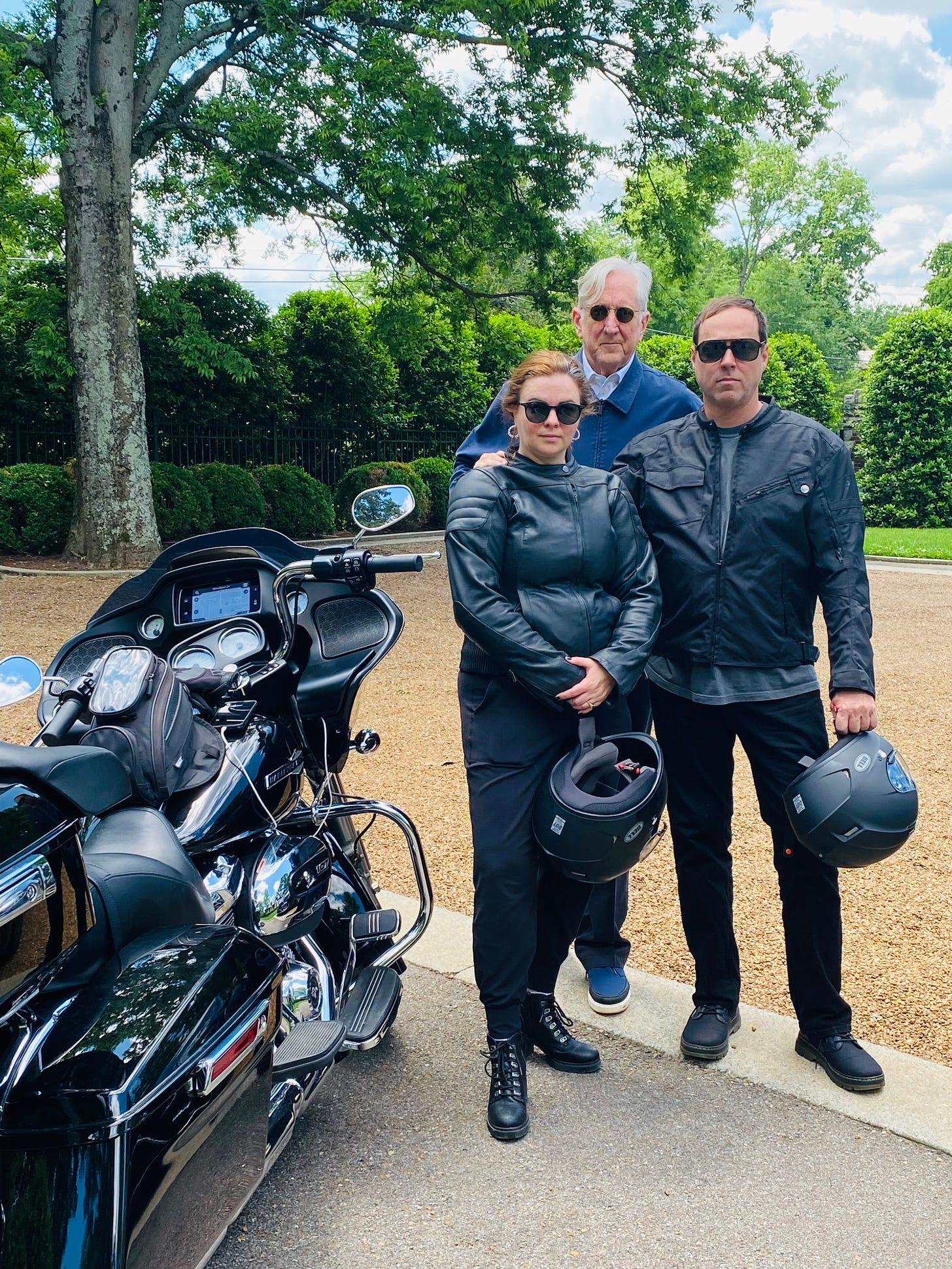 Amber and Derrick stand next to their parked Harley. They are wearing their riding gear and are holding their helmets. Behind them, T Bone Burnett stands with his hand on Amber's shoulder. They all wear sunglasses and look into the camera, unsmiling. 