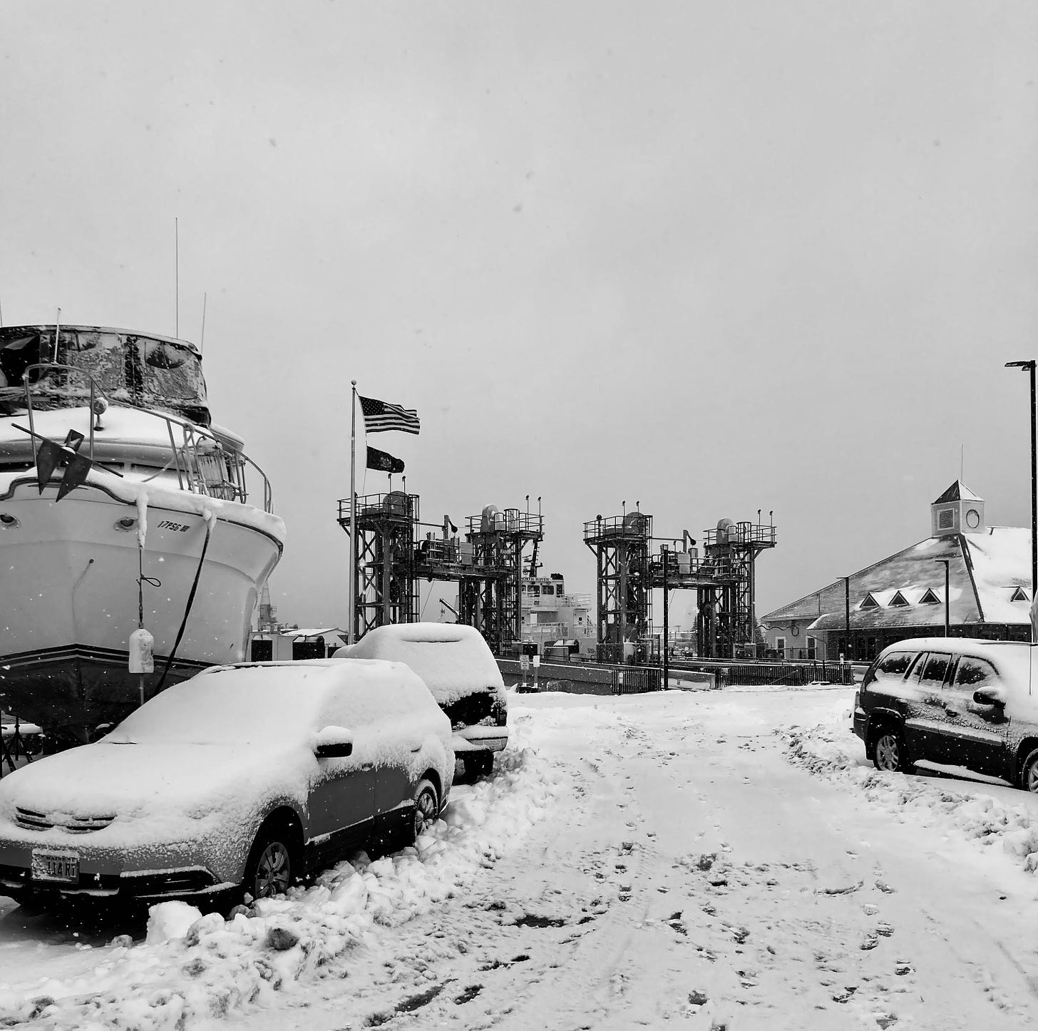 A snow scene in black and white. In the foreground are snow-covered cars and a boat, and beyond that a ferry terminal. 