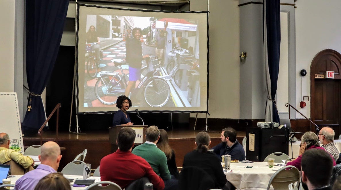A room of mostly white-male presenting people seated around tables watches Kristen, who is wearing a blue silky dress, big poofy hair, black cat-eye glasses and standing at a lectern with an image of her at a bikeshare demo in her hometown of Greensboro projected behind her head