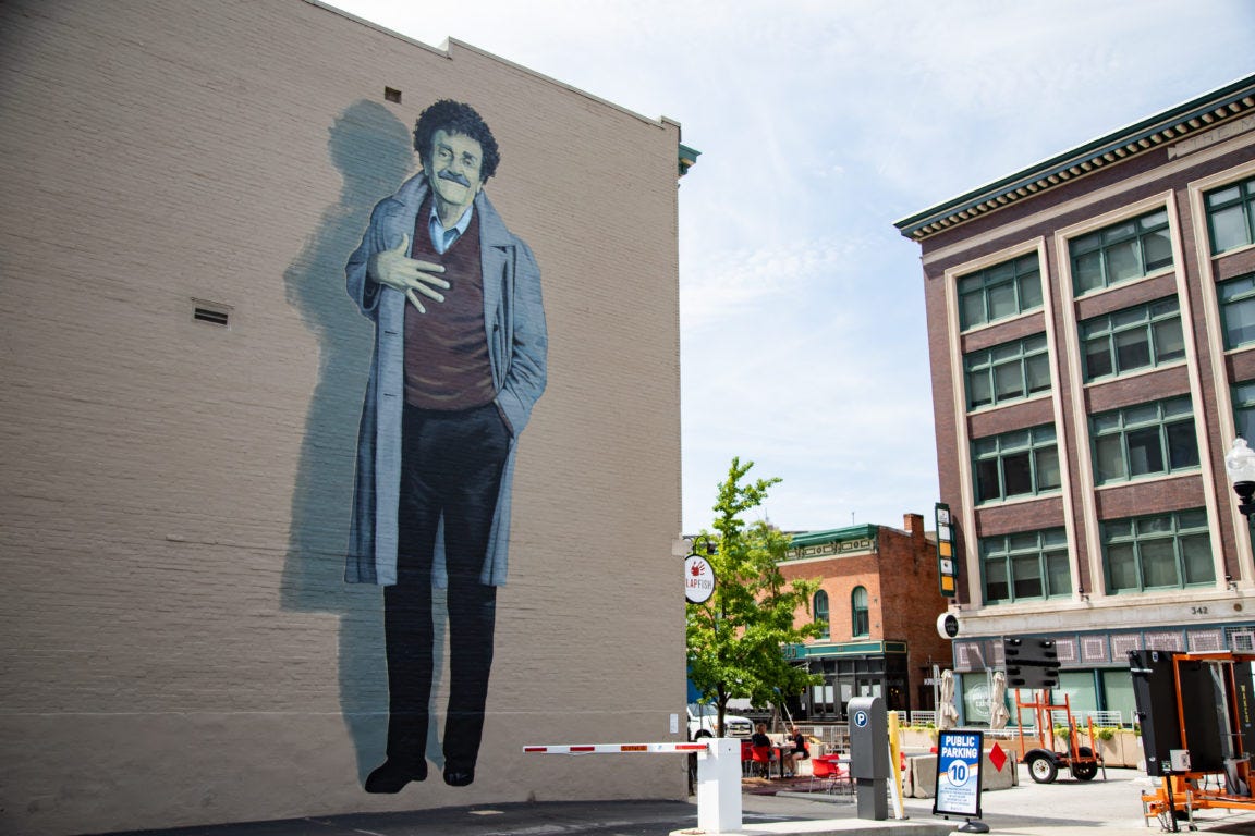 Four story mural of Kurt Vonnegut on the side of a building. Vonnegut stands with a long coat open, a hand on his chest, and his other hand in his pocket. He has a slight, wry smile.