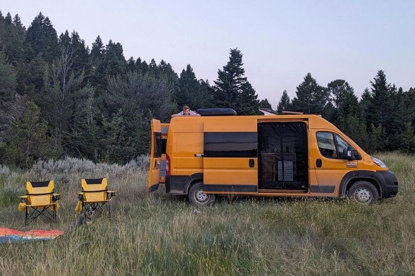 A bright yellow campervan sits in a grassy field to the right of two yellow lawn chairs and a rainbow-colored blanket. Next to the lawn chairs is a blue heeler hiding in the tall grass.