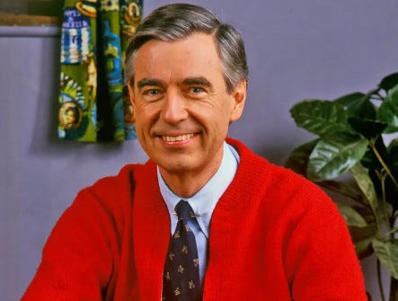 Mr. Rogers smiles into the camera. He is late-middle-aged with salt-and-pepper hair cut short, and a bright red cardigan sweater.