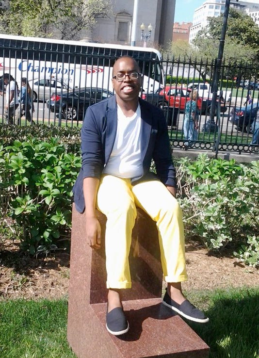 Picture of Black male sitting on statue outside, wearing a navy blazer, white t-shirt, yellow chino pants and black slip on shoes.