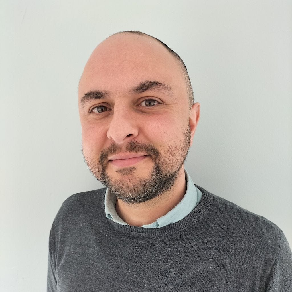 white man with shaven head and dark beard, wearing a grey jumper over a blue shirt