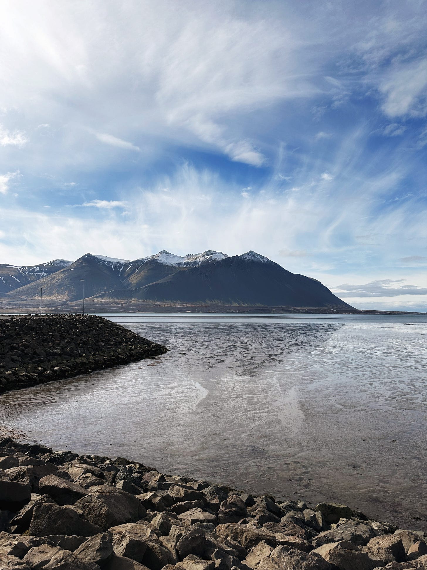 The view across the water to a snow capped mountain under bright blue skies with wispy white clouds in Borgarnes, Iceland.