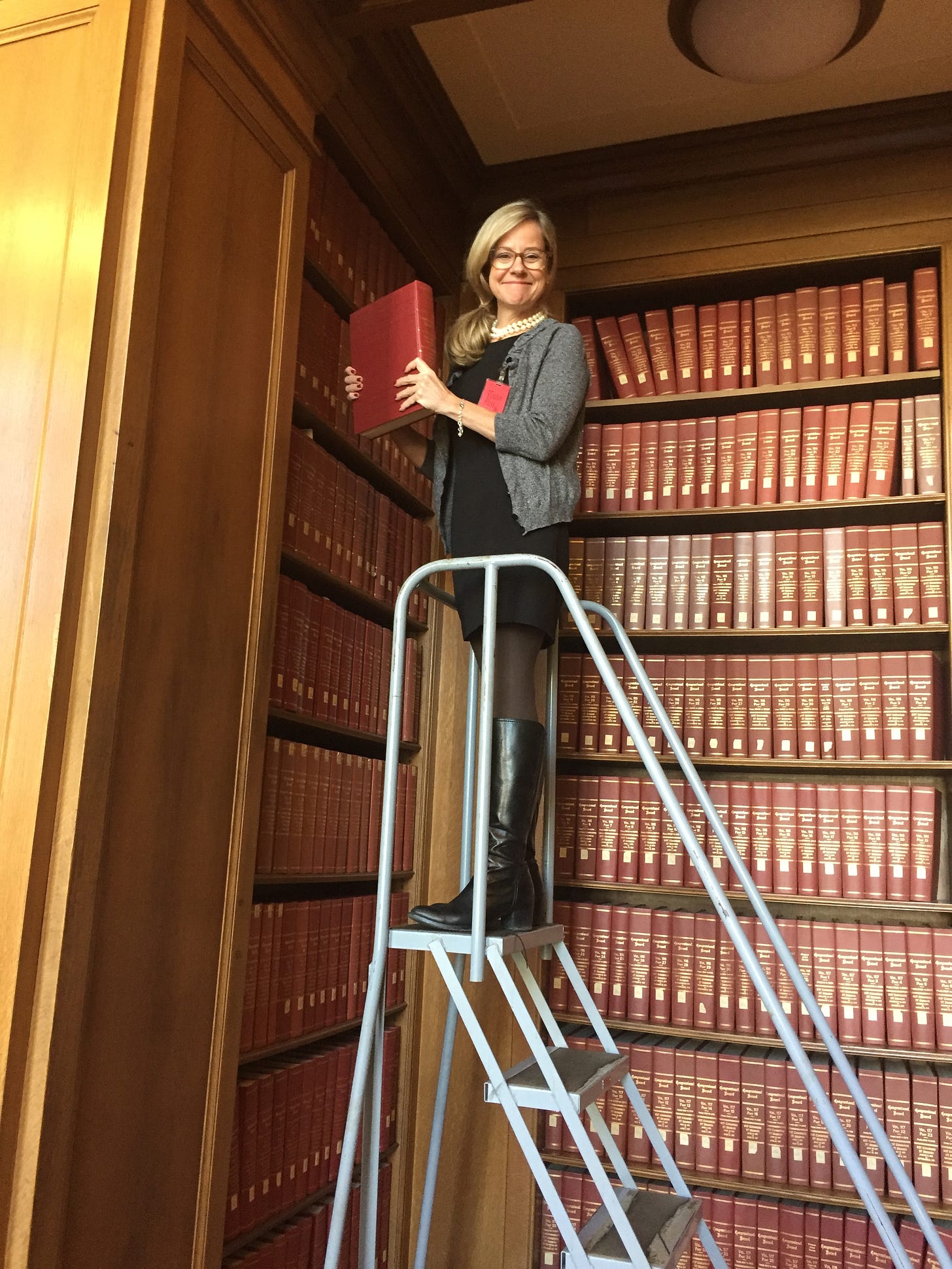 Author/speaker/memoir-writing coach Christine Wolf in Washington, D.C., conducting research in The National Archives (2017). She's wearing a gray cardigan, black dress and boots, and pearls. She's on a high ladder, holding a red book, surrounded by red archival volumes.