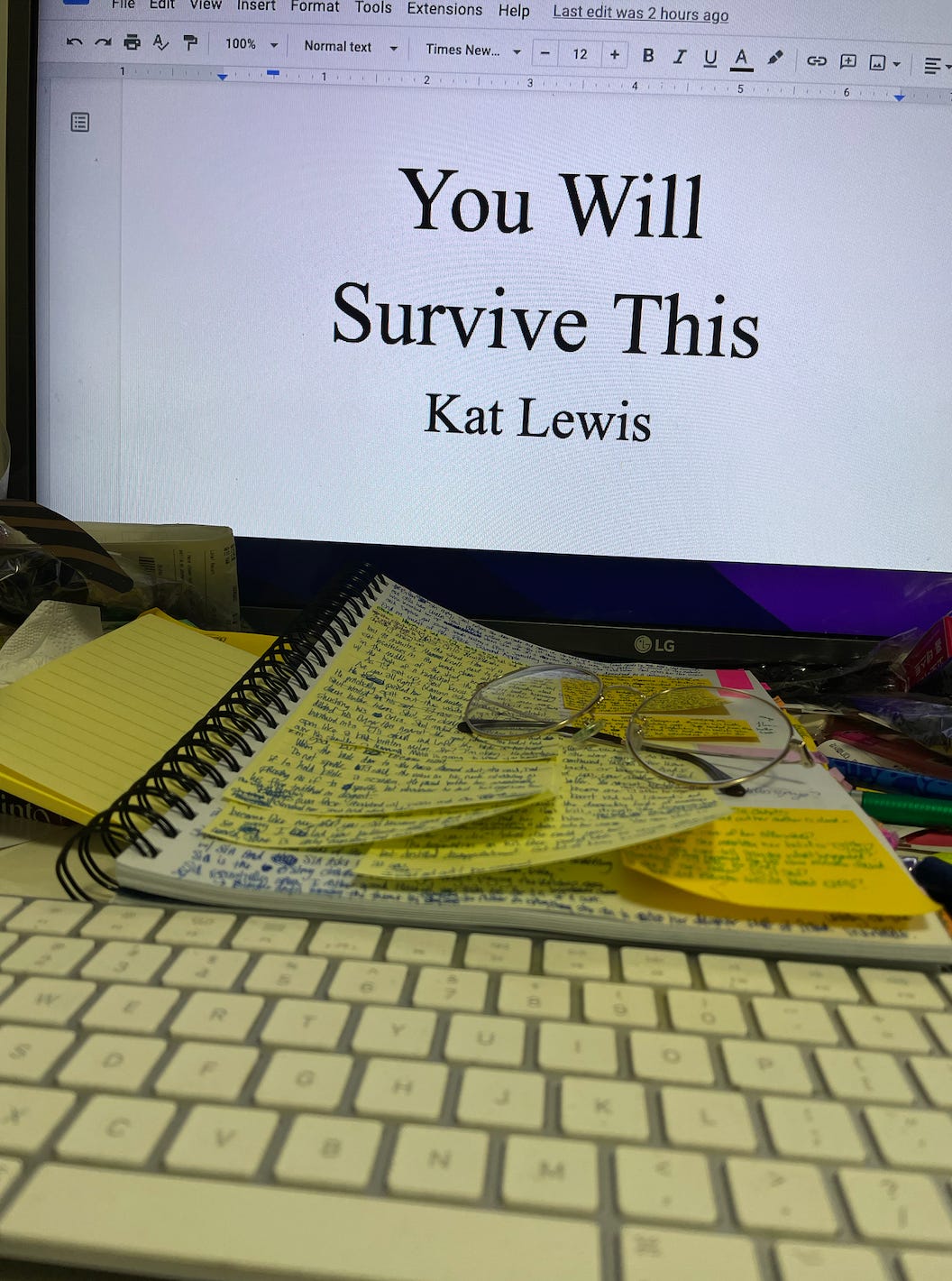 Kat's computer monitor is opened to the title page of her novel You Will Survive This. In front of the screen, her desk is messy with trash—wrappers, receipts, and scattered pens. Her printed manuscript rests between her keyboard and monitor. The page is covered in several post-it notes of difference colors and sizes. Messy hand writing covers every inch of the page and post-it notes.