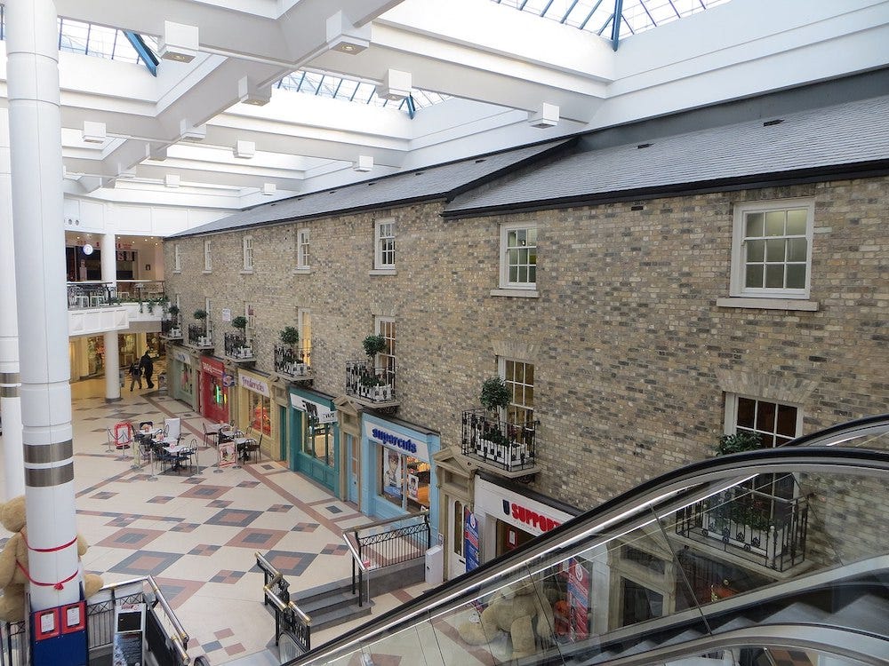 Marlands shopping centre in Southampton. This is a view of the interior, with an escalator going down to the ground floor next to a fake Georgian street which features various shopfront and above them panelled windows with balconies and fake trees upon them – outside the shops there are some tables where you can sit and have a drink. There are barely any people in the shopping centre
