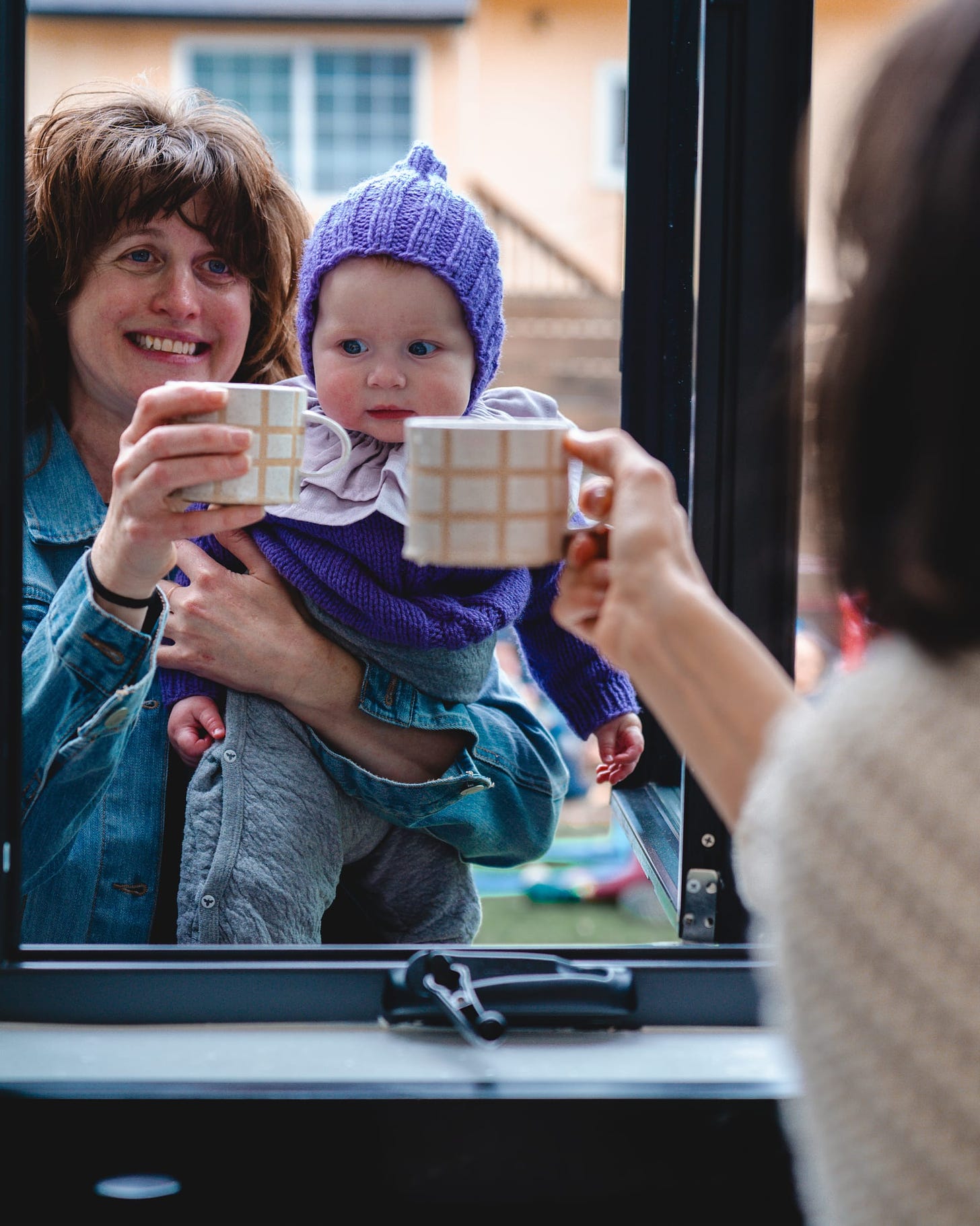 someone in a denim jacket holding a baby and clinking mugs through an open window