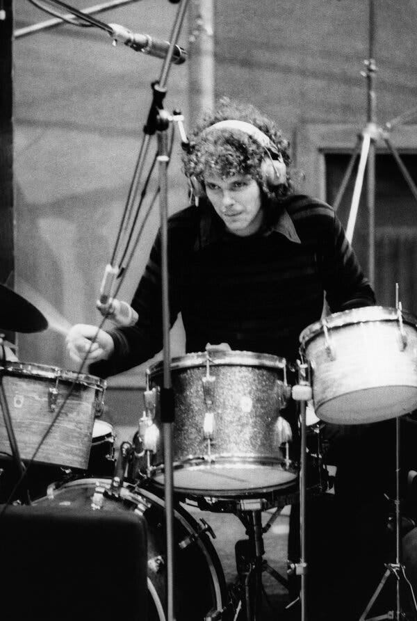 Jim Gordon, an intense-looking young man with a big mop of curly hair, playing the drums in a studio. He is wearing dark clothes and has headphones on.