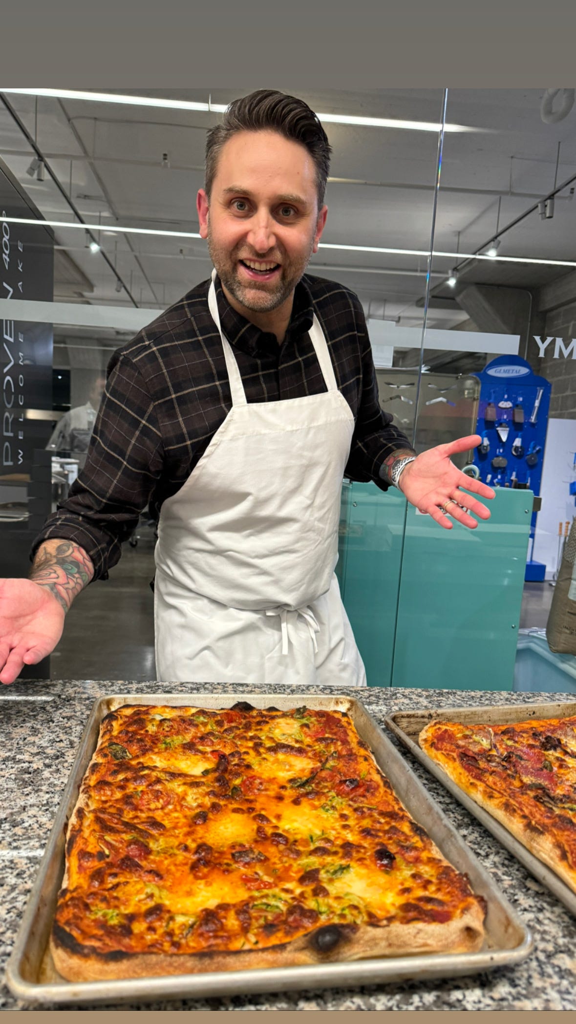 My friend Matt at pizza class. He's wearing and apron and posing proudly with his freshly-baked Pizza Romana, which is on a sheet pan on the counter in front of him.