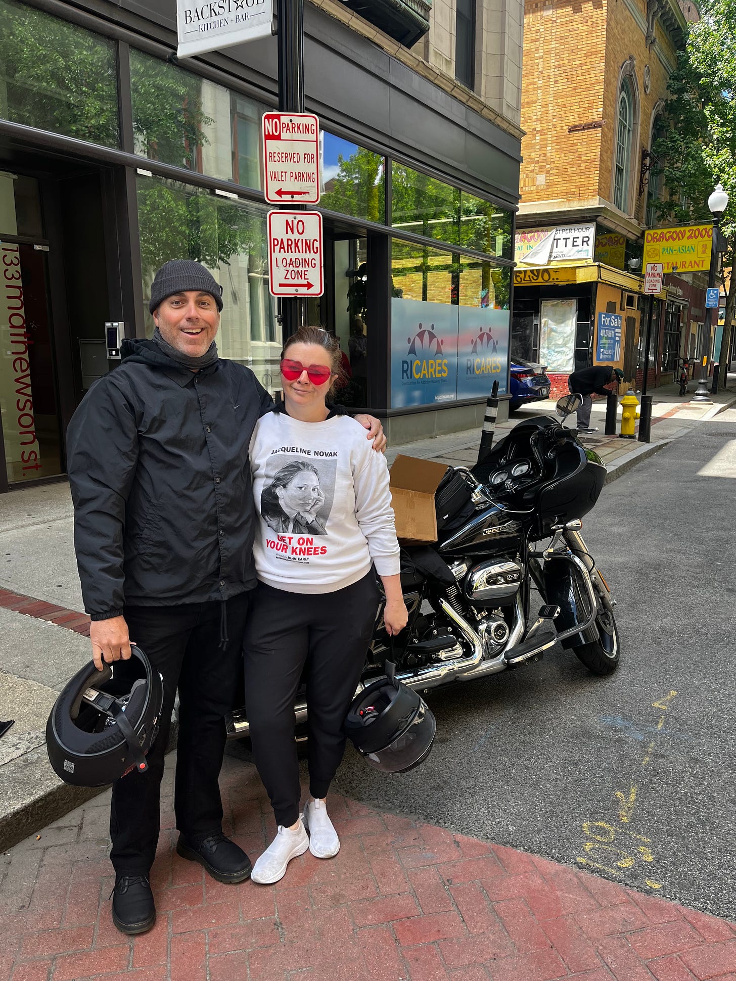 Derrick and Amber pose with their arms around each other in front of their parked motorcycle. Amber wears red heart-shaped glasses and smiles slightly. Derrick holds his bike helmet and is smiling, looking excited.