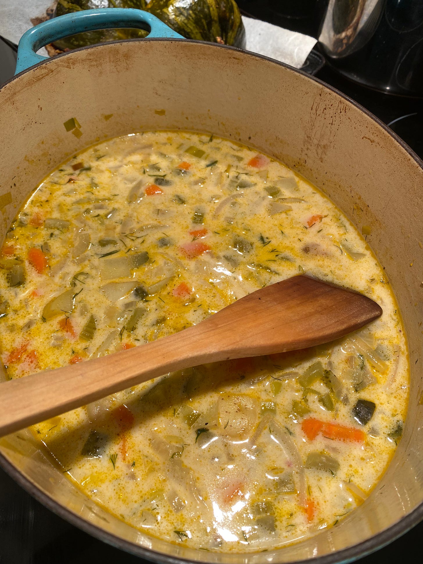 The pickle soup described above, a creamy yellow broth with bits of carrots, pickle, onion, and celery, in a blue oval dutch oven with a flat wooden spoon in it.