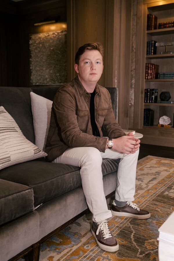 Thomas Fleming, wearing light-colored pants, a brown jacket, brown shoes and a shiny watch, sits on a brown sofa, a bookshelf behind him.