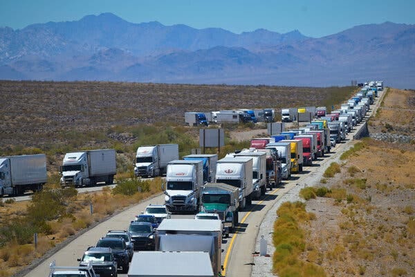 A view of a long line of trucks and cars closely spaced on a highway in an open area. Mountains rise in the background.