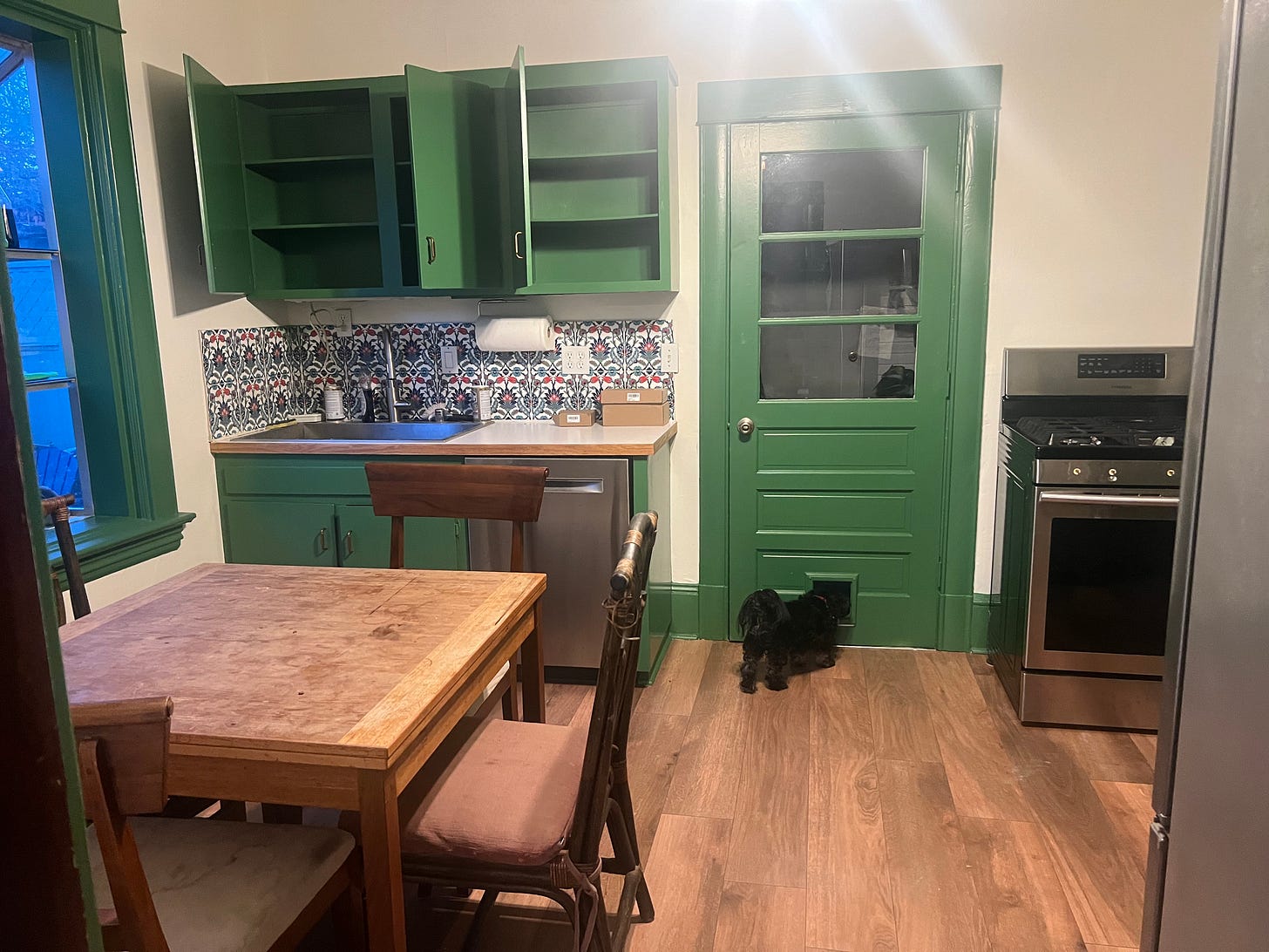 Kitchen with blue patterned tiles under some green cupboards. Also a wooden table and chairs.