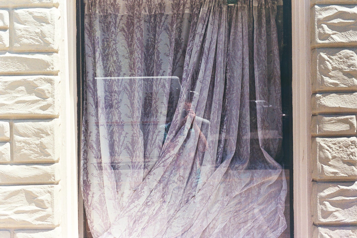 a self portrait in the reflection of an exterior window obscured on the inside with wrinkled floral drapes