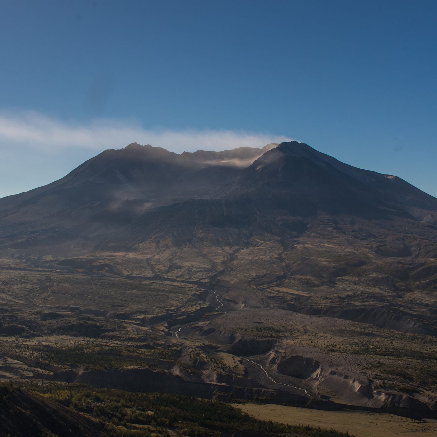 Bare slopes and crater of Mt. St. Helens with white smoke or cloud blowing from it under a clear blue sky.