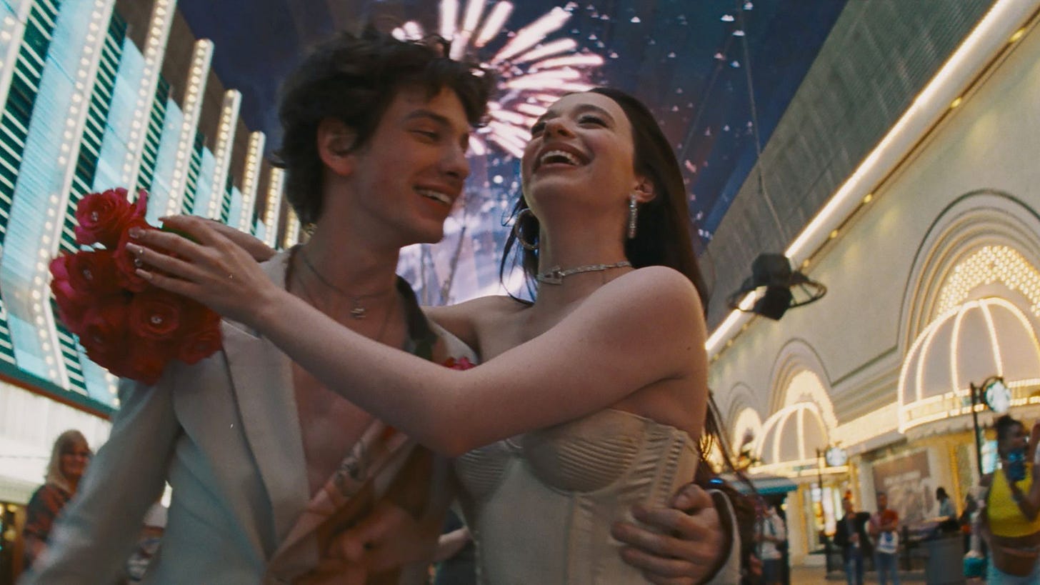 A low angle shot of Vanya (Mark Eydelshteyn) embracing Anora (Mikey Madison) against a backdrop of fireworks