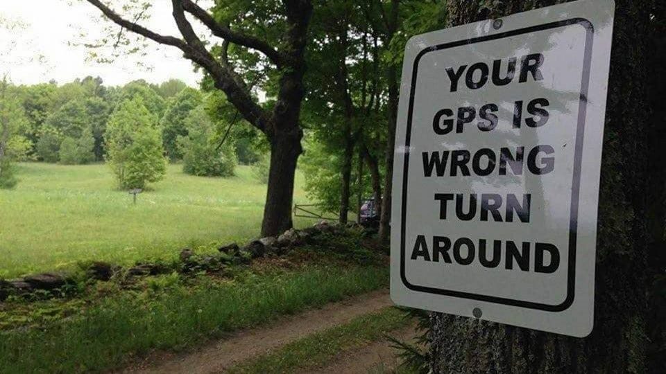 A sign posted on a tree near a dirt road adjacent to a grassy field reads: "YOUR GPS IS WRONG. TURN AROUND."