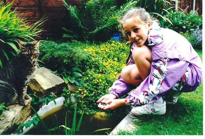 a girl holding a baby frog at a pond with a cat looking on