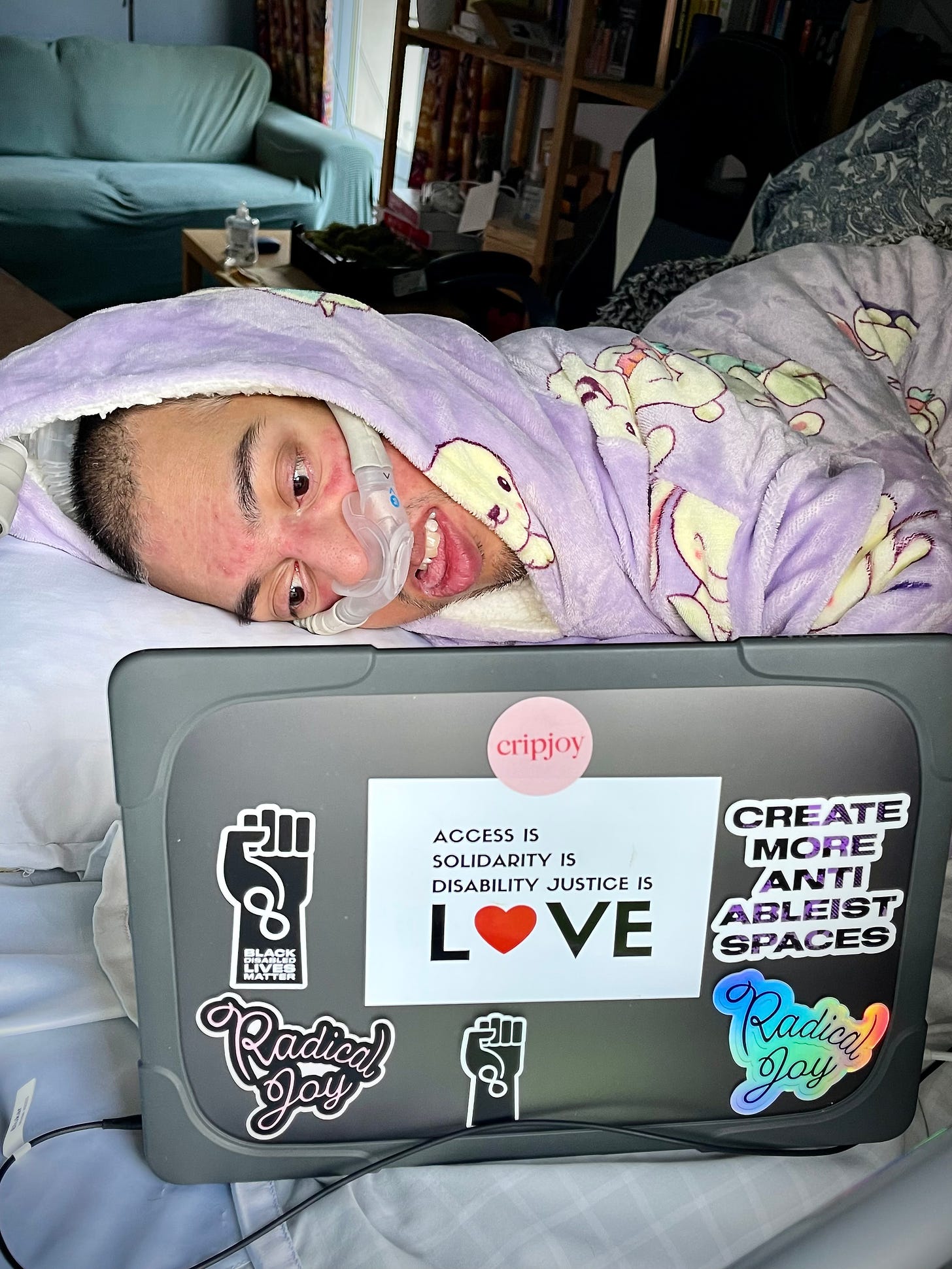 Image description: Bold, wild, fearless existence. In the middle of an abstract dark blue background is a photo of Sulaiman (a wholeheartedly Disabled AF and British-Pakistani man) lying in bed on his right with a nasal ventilator mask over his face as he works on his MacBook Pro laptop. On the grey case of his MacBook Pro are 7 stickers on the back of the screen via Sulaiman himself and his Disabled global kin (by bond) – Jennifer White-Johnson, Alice Wong, Sandy Ho, and Mia Mingus. Sticker 1 is a circular pink sticker with red text that says “Cripjoy.” Sticker 2 is a sticker with a white background featuring a gradient black to purple text that says “Create More Anti-Ableist Spaces” in capital letters. Stickers 3 and 4 are of cursive text that says “Radical Joy,” one a sticker of pink text with a black outline and the other a colourful holographic sticker. Sticker 5 is a graphic of the black power fist against a white background with the infinity symbol and the words “Black Disabled Lives Matter” written in bold white font placed down the wrist of the fist. Sticker 6 is a graphic of the black power fist against a white background with the infinity symbol for neurodiversity in bold white lines placed down the wrist of the fist. Finally, sticker 7 is an extensive rectangular white background with black text over 4 lines; on lines 1 to 3, the text says, “Access is… Solidarity is… Disability Justice is…” and on line 4, the larger text says, “Love” in capital letters with the “o” replaced with a red heart emoji. Sulaiman is wearing his Bunny Oodie (“like a jumper crossed with a blanket crossed with a cloud”) in purple via @the_oodie, and he feels calm as he works on his MacBook Pro. At the top middle of the image is cursive white text that says, “Sulaiman R. Khan.” Below it, in all caps, white text is “Daringly Integrating Disability.” At the bottom right corner of the image is the social logo of ThisAbility Limited in all white featuring the Adinkra symbol for interdependence. End.