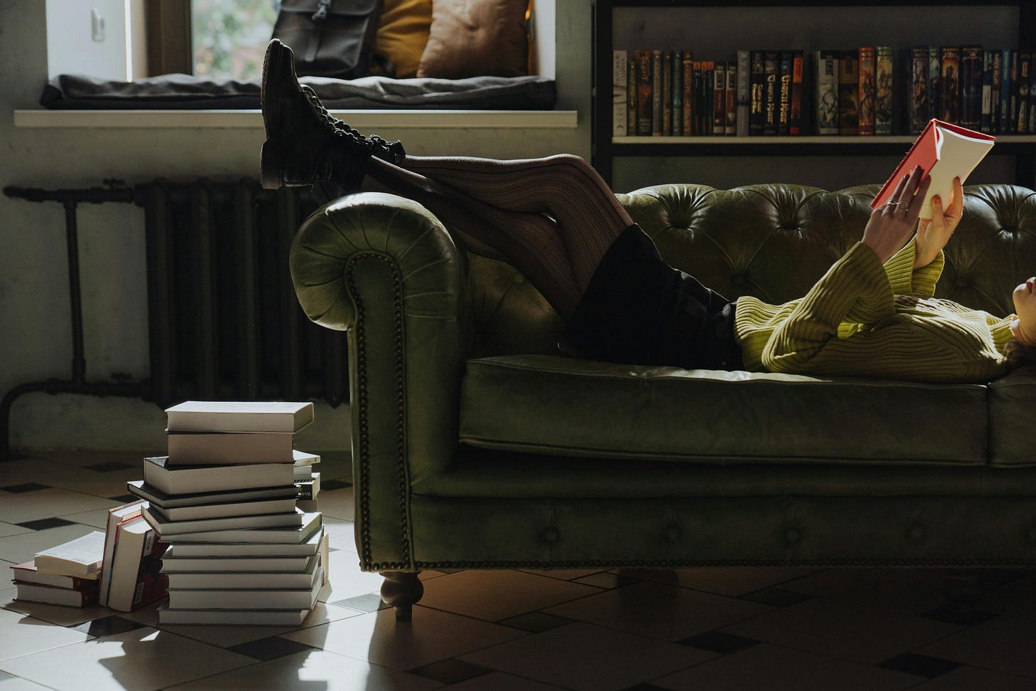 girl wearing a green sweater lying on a leather couch reading a book with a red cover