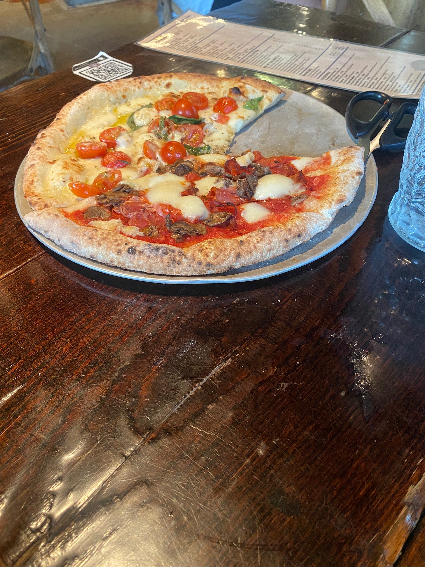 On a wooden table, a thin metal serving plate holds leftovers of two pizzas: one, a tomato base with roasted mushrooms and salami, the other, an oil base with cherry tomatoes and basil. Both have little melted dollops of fior di latte. A pair of pizza scissors are visible partially tucked under the tomato sauce pizza.