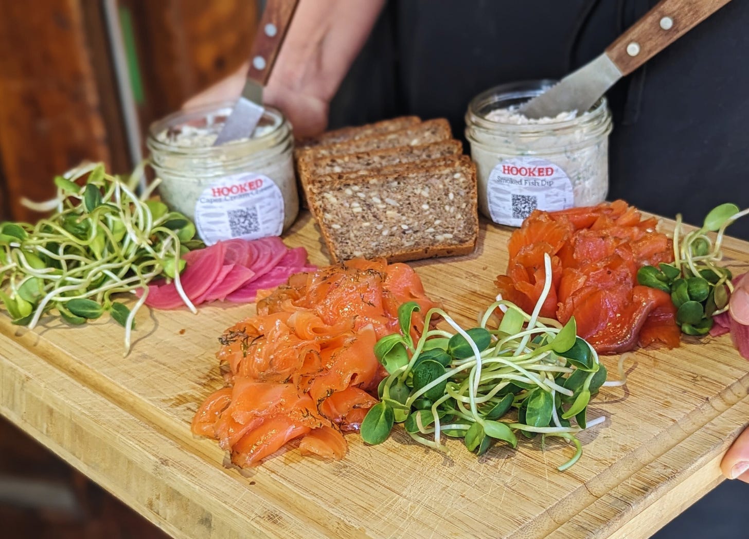 hands hold a wood board with piles of artfully arranged fixings for smoked fish: three piles of sunflower greens, two arranged servings of smoked fish with some dill sprinkles, two jars with cream cheese and smoked fish dip, two servings of sliced pickled beets and homemade rye bread.