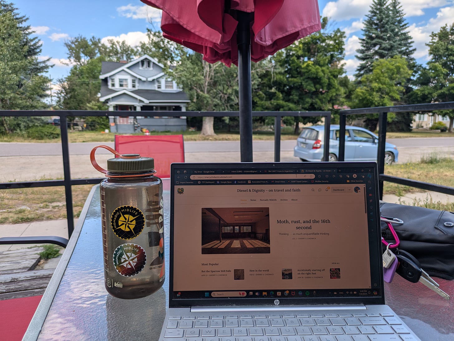 a laptop and water bottle on a patio table in t he shade with a small car and a historic home in the background, against a blue sky with puffy clouds