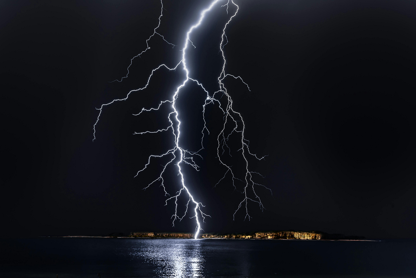 A complex lightning bolt strikes the open ocean in front of some lights. The background is pitch black, so the lightning is crisp, and is bright white.