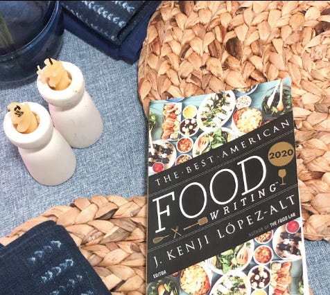 overhead photo of woven placemats, yellow candles in white holders, blue napkins and a book that reads “The Best American Food Writing” and “Kenji Lopez-Alt”