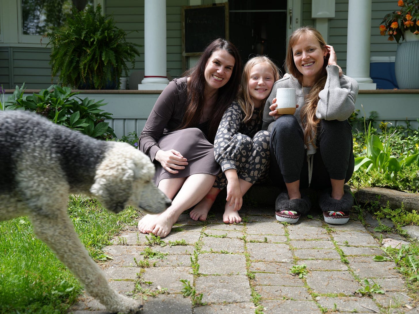 Three people sit on a stone walkway in front of a house, there are two adult women with a little girl sitting in between them. They smile at the camera, and there is a dog walking into the frame from the left. Three people sit on a stone walkway in front of a house, there are two adult women with a little girl sitting in between them. They smile at the camera, and there is a dog walking into the frame from the left.