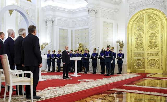 A man stands behind a podium flanked on each side by rows of soldiers and citizens. A massive golden door is beside him.