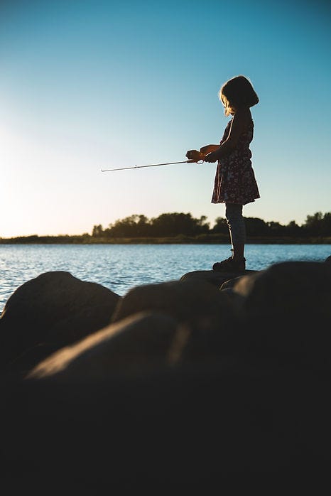 Photo of a little girl standing on rocks, holding a fishing rod while looking out to the water. 