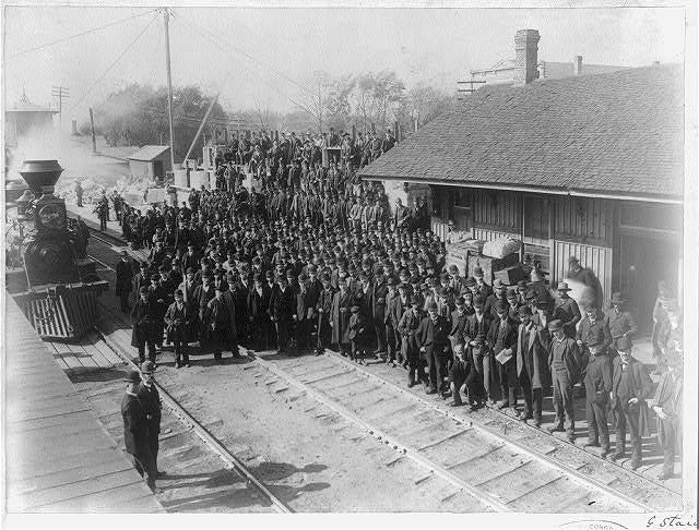 Photograph shows a large crowd of men gathered on railroad platform and tracks; train in background on left. 