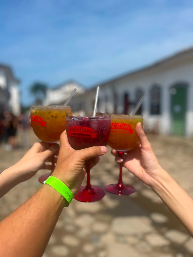 três mãos em close brindando com taças grandes de drinques coloridos. Ao fundo, se vê ruas de uma cidade histórica num dia ensolarado fora de foco.