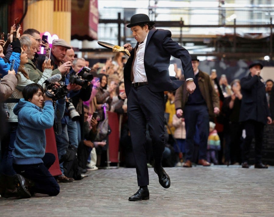 People watch as a competitor wearing a suit and bowler hat runs awkwardly while carrying a pancake on a frying pan.