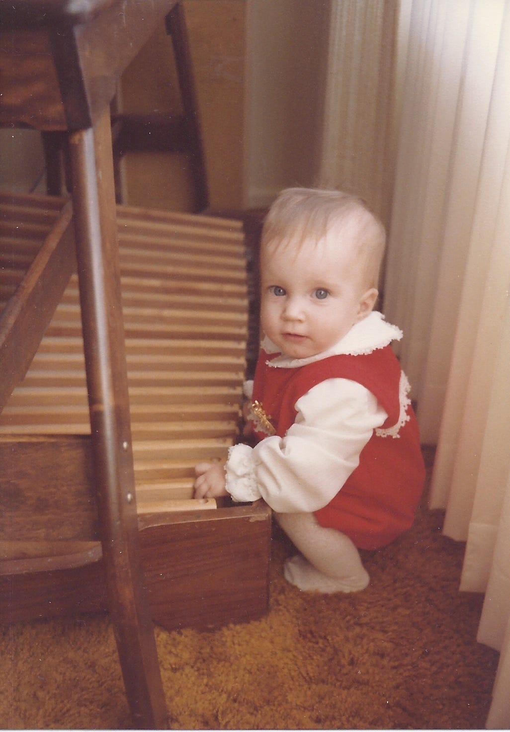 image of the author as a baby, wearing a red outfit and squatting next to the pedals of an organ