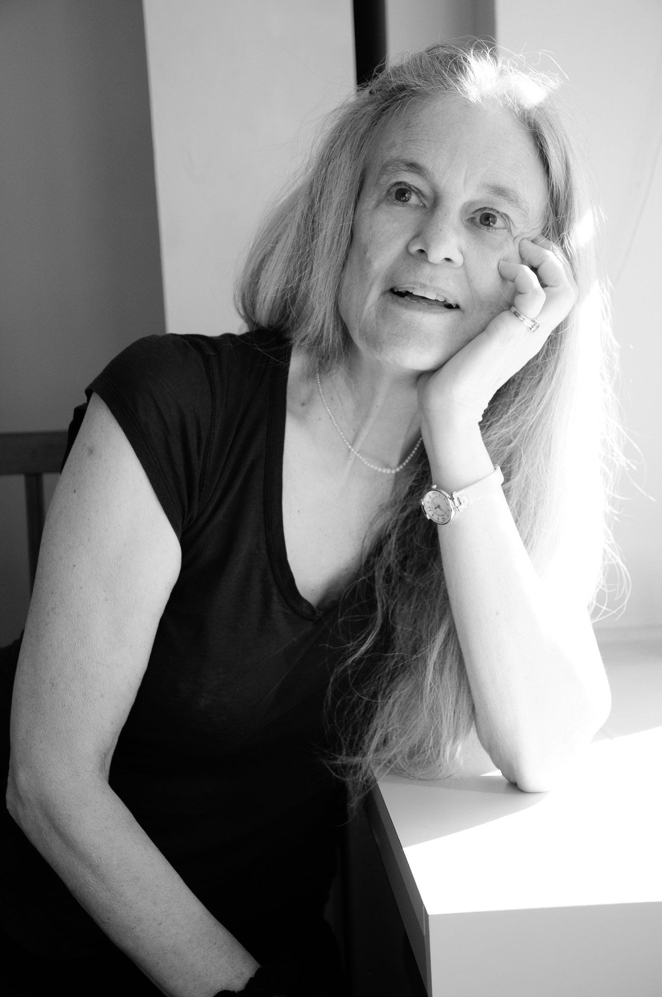 A recent black and white photo of Sharon Olds from the waist up. Her elbow is on the table and her hand props up her chin. She looks up and to her right. Her lips are parted as if she's speaking to someone.