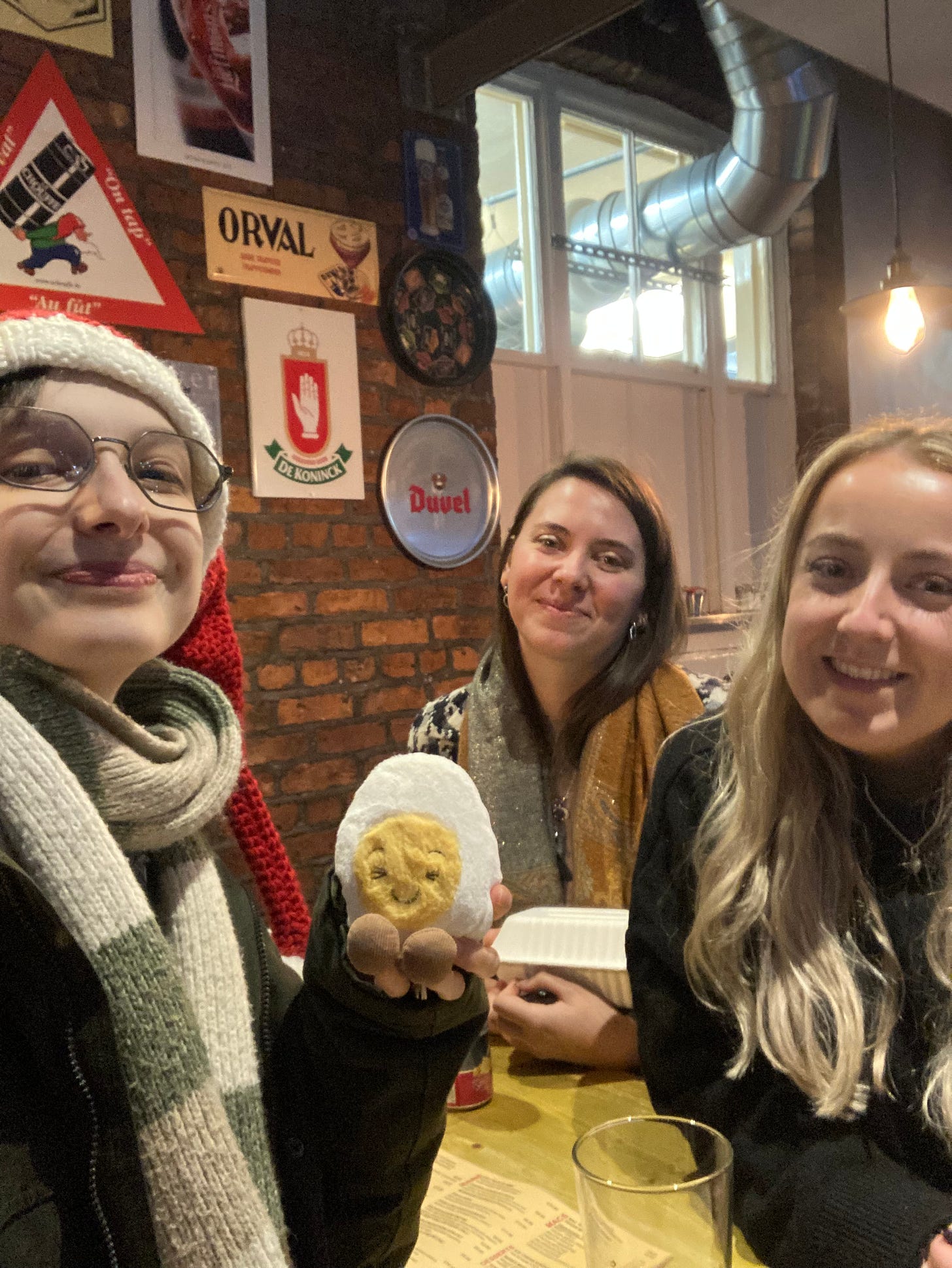 Selfie of three white PhD students smiling around a bench table in a pub with exposed brickwork and metal signs hanging on the wall. Dippy the soft toy egg is in the middle held by Beck who is wearing a knitted santa hat. 