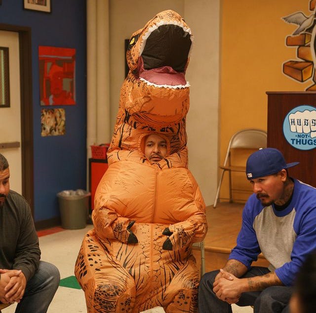 A man in an inflatable t-rex costume sitting in a therapy session with two other men of color. 