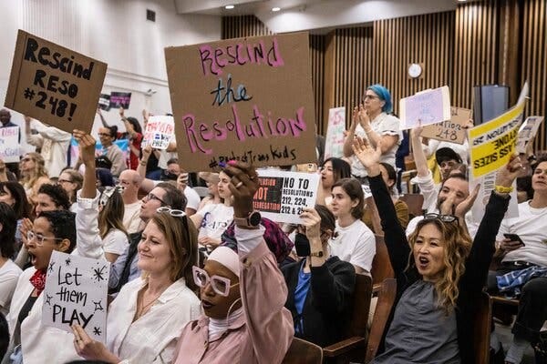 A roomful of seated parents smile and hold up signs, including one that says “Rescind the resolution.”