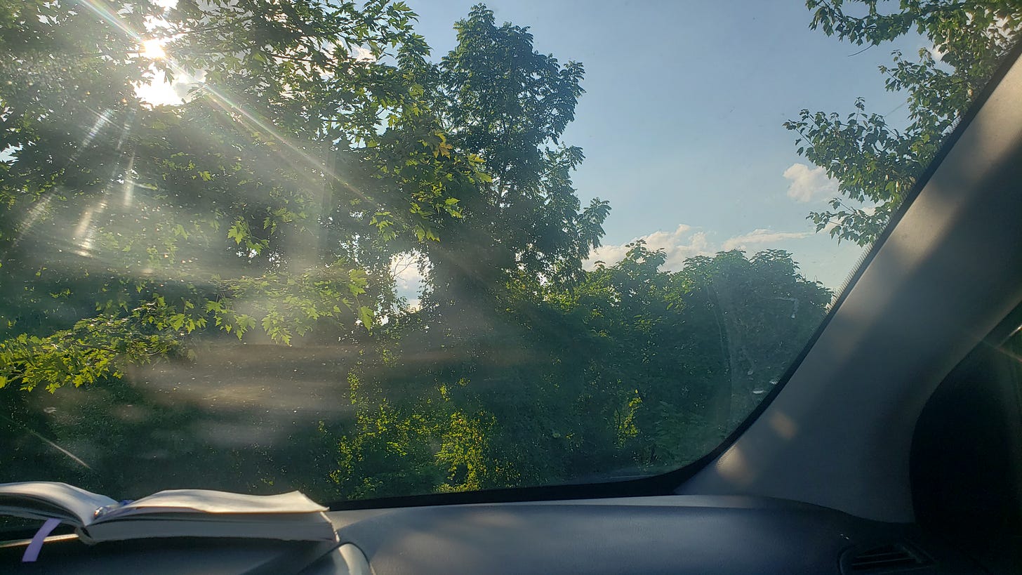 Picture taken through front car windshield of green trees with rays of sun shining through the leaves, and blue sky. An open notebook with a pen is perched on the dashboard..
