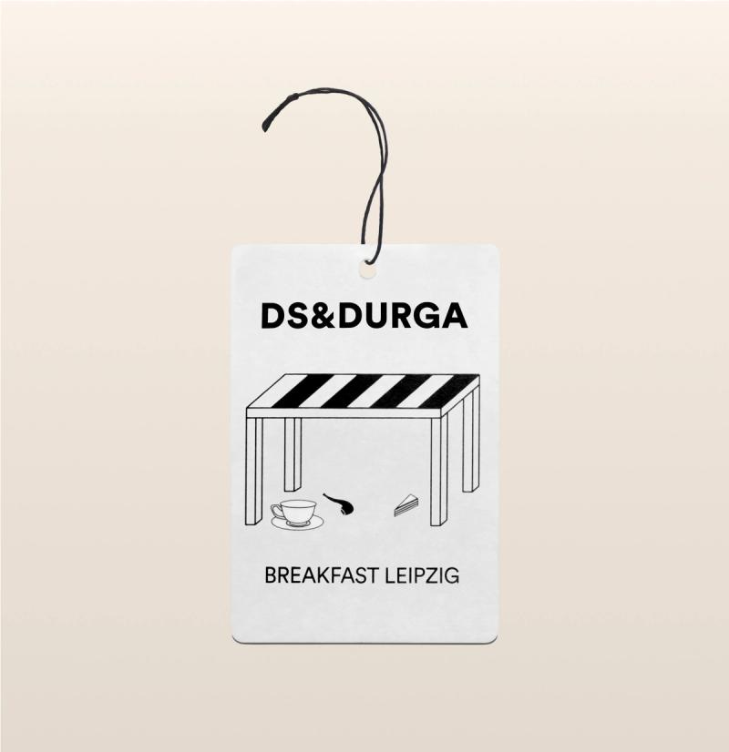 A hanging auto fragrance with the inscription"ds & durga" on the upper portion and "breakfast leipzig" below featuring stylish monochrome design with a table, cup, and spoon illustration.