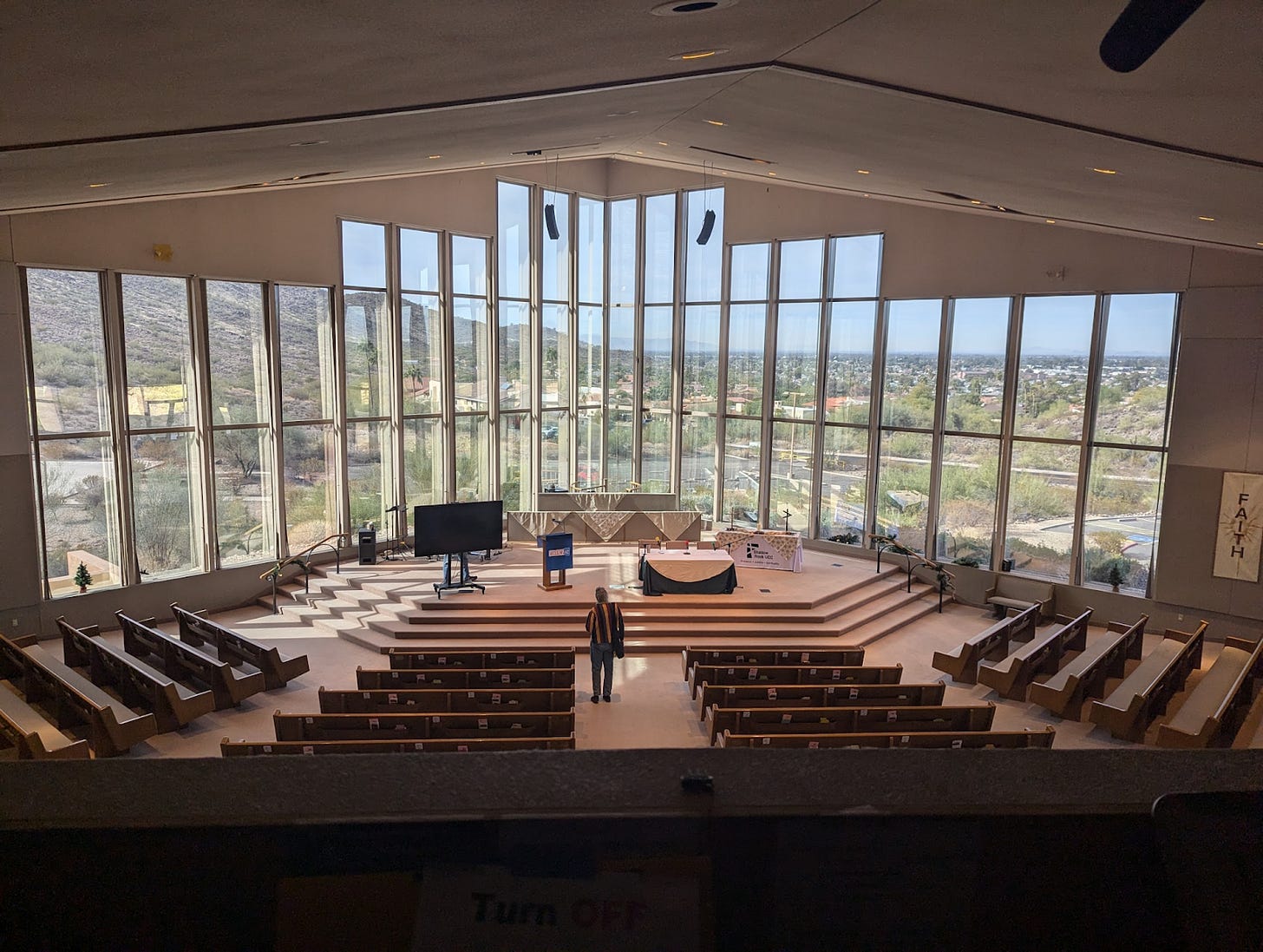 Photo taken in the sancutary at Shadow Rock UCC. There are rows of pews leading toward a wall of windows that overlooks a stunning view of the desert beyond. A lone figure stands near the front Dias.