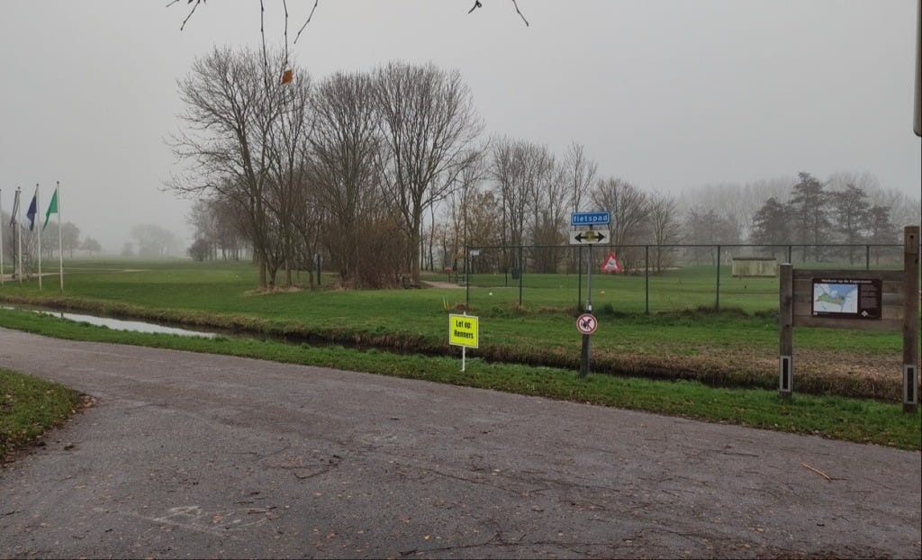 Dutch watch out for runners sign, early on in the loop, with a canal and green grass behind the paved path, and tall spindly trees dotting the golf course.