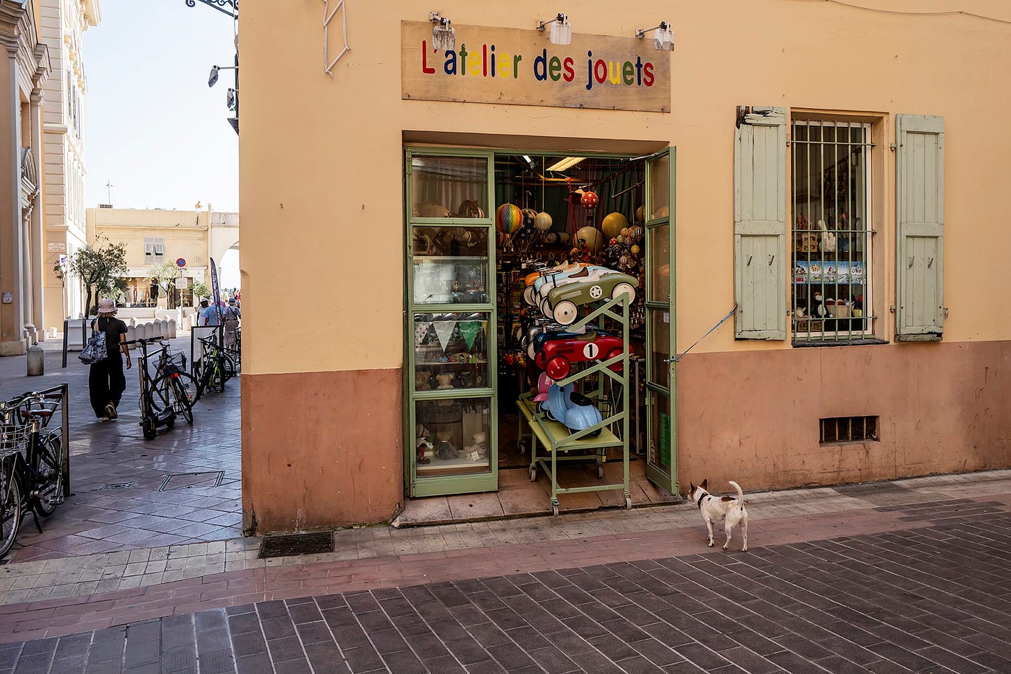 Vintage Toy Shop in Nice – A charming storefront with the sign “L’air des jouets,” featuring a colorful window display of toys. A small white dog stands on the sidewalk outside the shop.