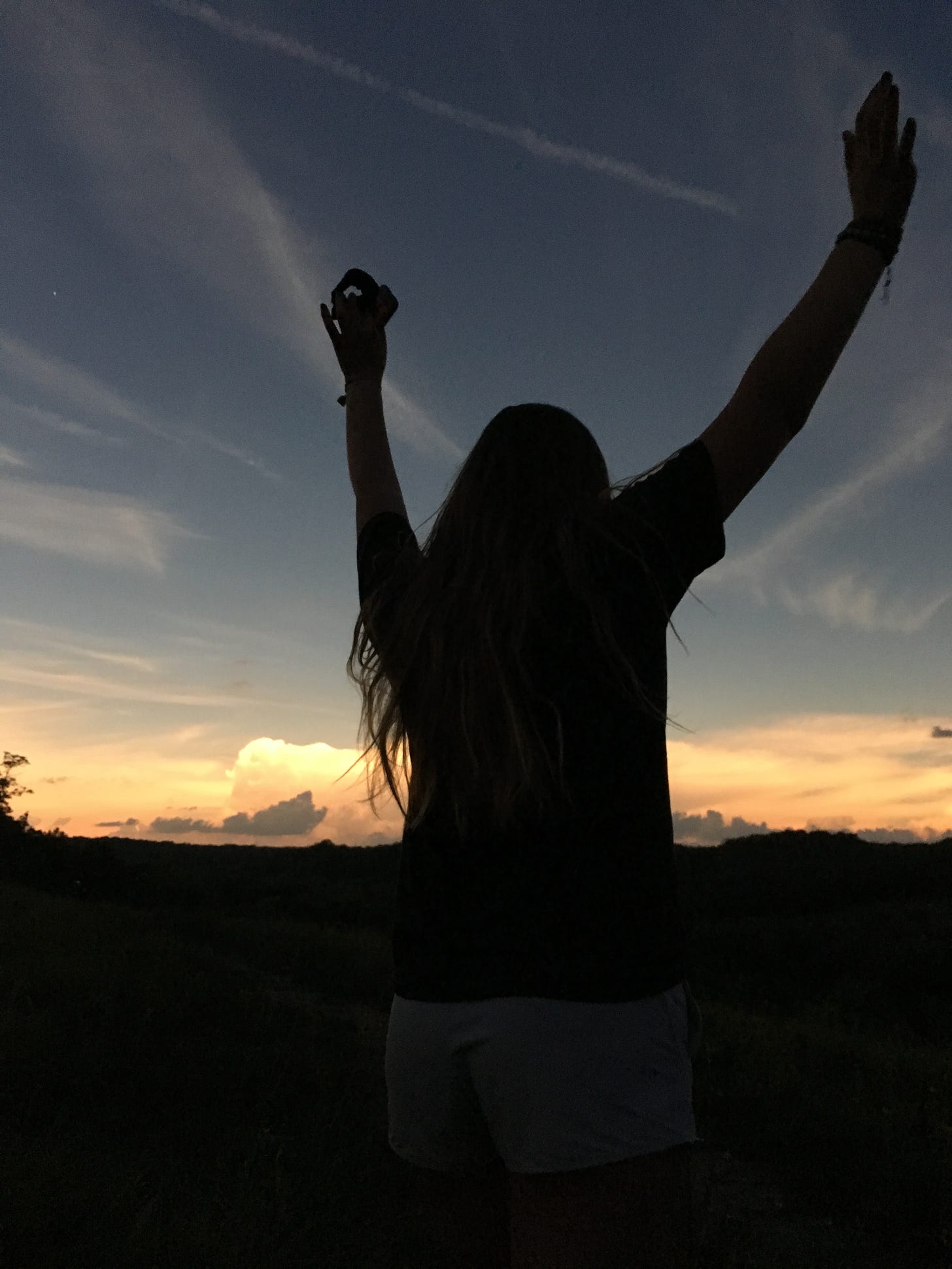 Silhouette of person with arms raised beneath a darkened solar-eclipse sky with the lit clouds on the horizon.